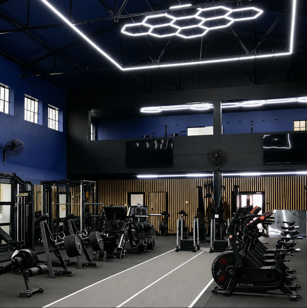 A modern gym interior with black exercise equipment, a dark blue ceiling, and large white hexagonal LED ceiling lights. The space includes multiple weight machines, a rowing machine, and a row of stationary bikes on a dark floor.